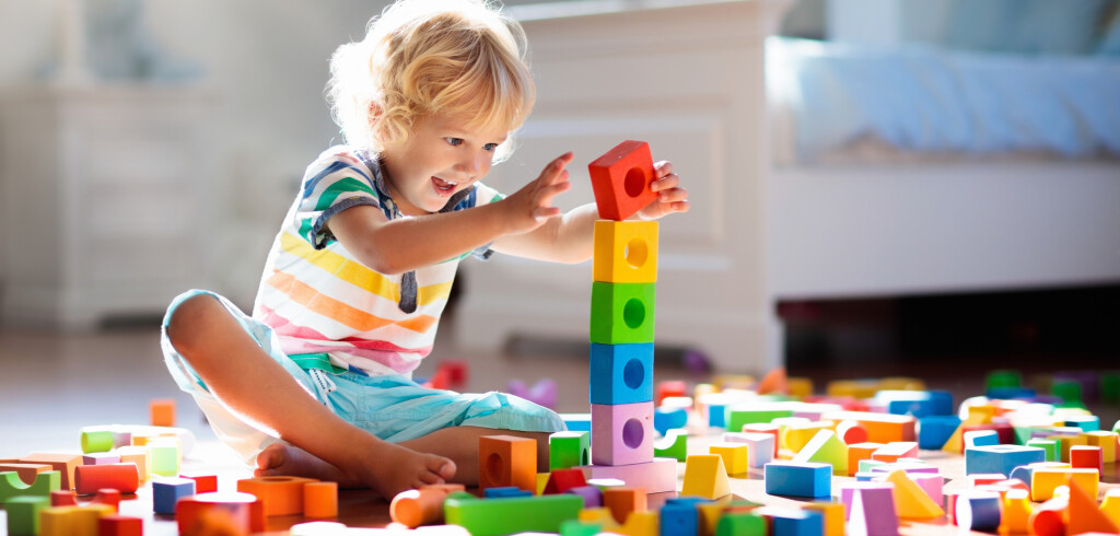 Child playing with colorful toy blocks. Kids play. Little boy building tower of block toys sitting on dark floor in sunny white bedroom. Educational game for baby and toddler. Children build toy house